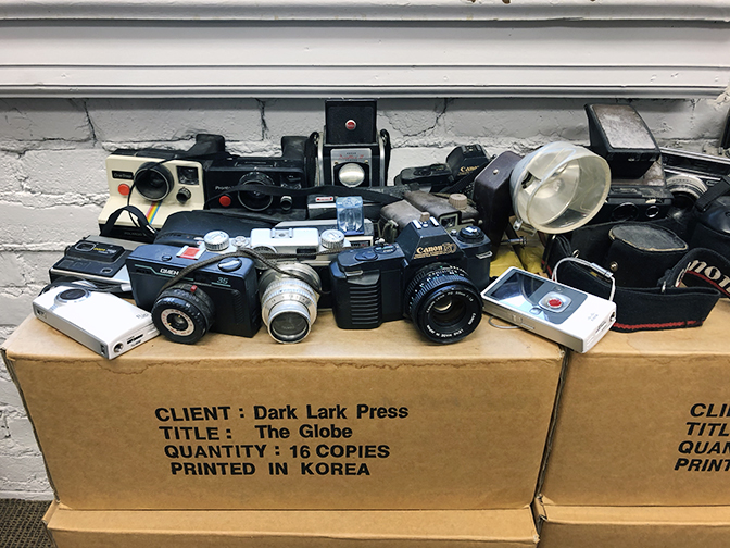 A collection of antique and digital cameras (still functioning) sit atop a box of "The Globe books".
